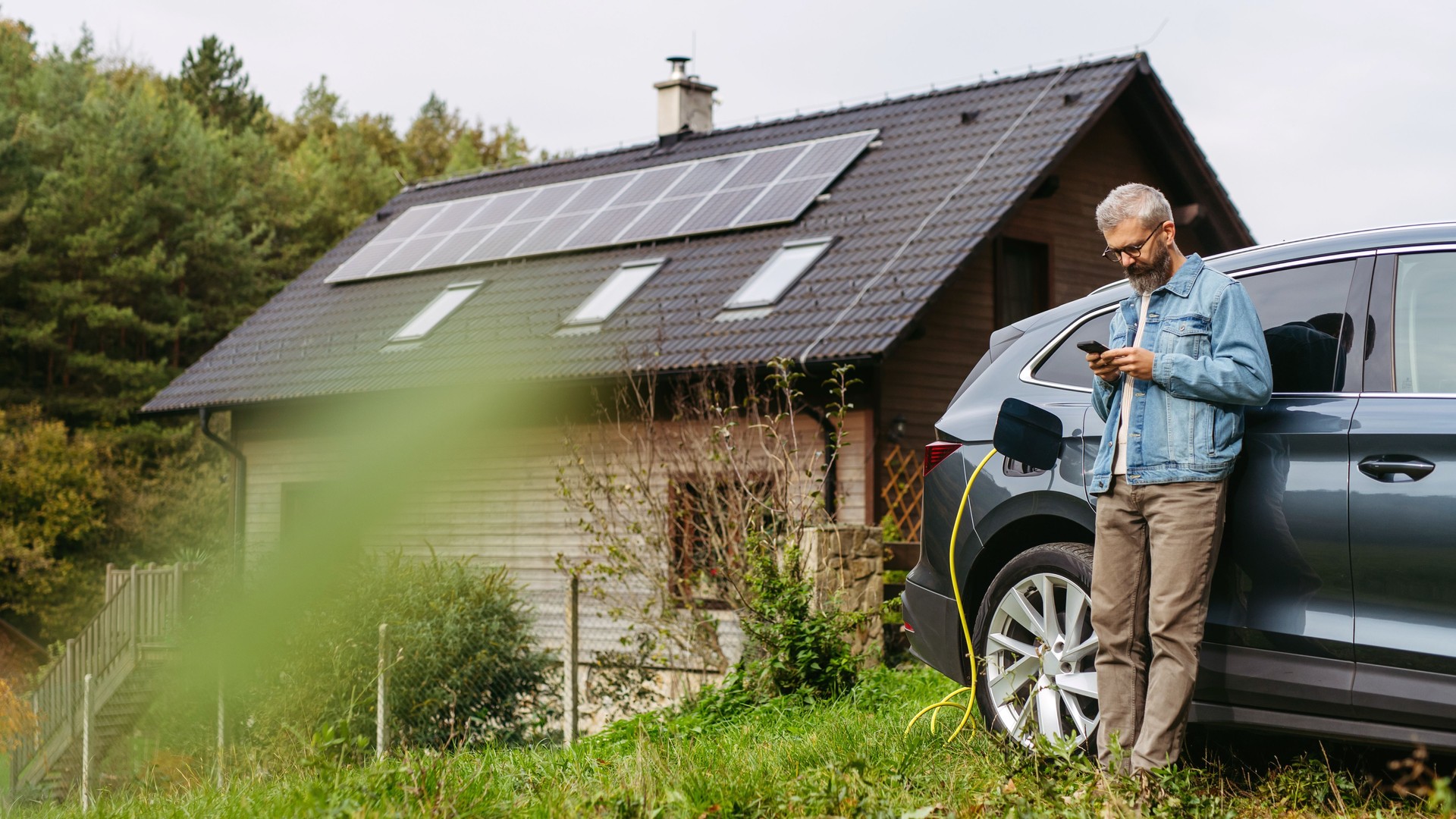 Man charging electric car in front of his house with solar panel system on roof behind him. Man leaning against a car while scrolling on smartphone.
