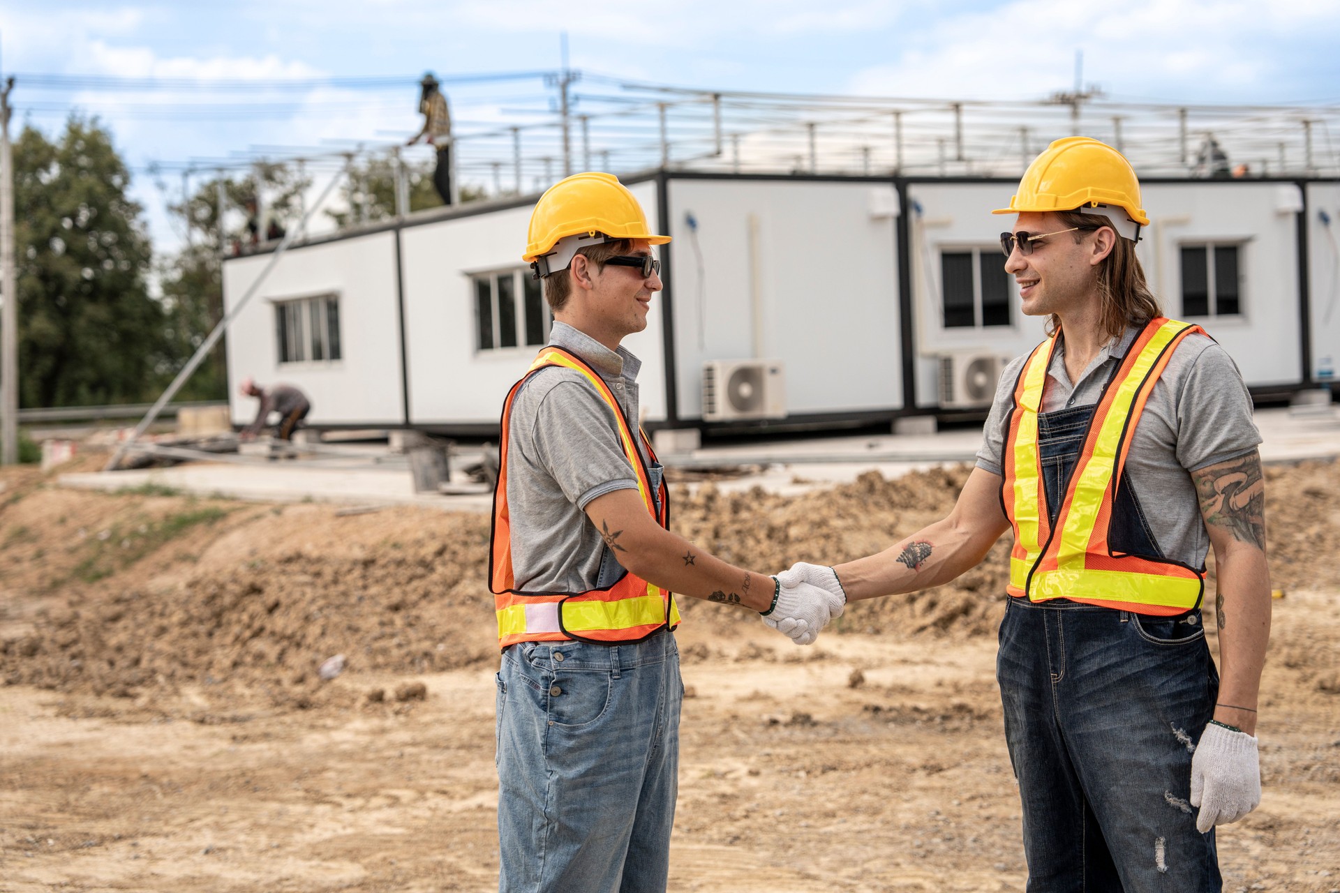 Two construction workers wearing hard hats and safety vests shaking hands at a construction site, symbolizing collaboration and successful teamwork with modular units in the background.